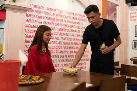 Waiter bringing a slice of pizza to a young Hispanic woman dressed in red. Waiter serving a girl at...