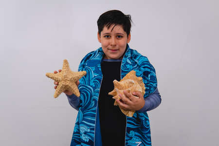A boy on a white background in a bathing suit, a towel, a seashell and a starfish in his hand. Boy...