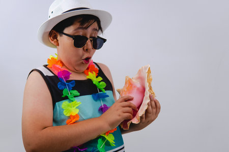 There is a boy on an isolated white background wearing sunglasses and holding a seashell. Boy with beach clothes, he has a sleeveless shirt, sunglasses, flower necklace and white hat and is holding a seashell.の写真素材