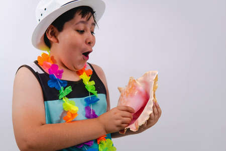 A boy on the beach dressed in a sleeveless shirt, white hat, and flower necklace, holding a shell...