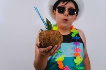 Boy dressed in beach clothes holding a coconut in the foreground. Boy wearing blue striped tank top, flower necklace, white hat and posing with a cocktail drink in a coconut.の写真素材