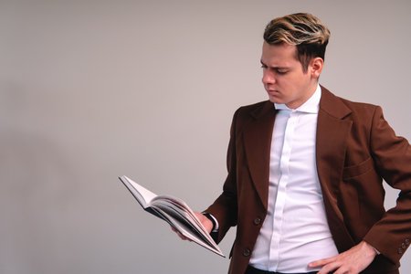 A blonde Caucasian man dressed in a brown jacket and white shirt with his hand on his waist reading...