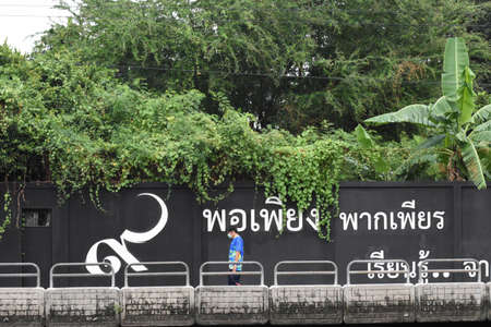 A man wears hygienic mask and walked on the walk way saen behide black wall with writting about no.9 (Thai Numeral) and philosophy explain in thai about Sufficiency economy at saen saeb canal Bangkok Thailandのeditorial素材