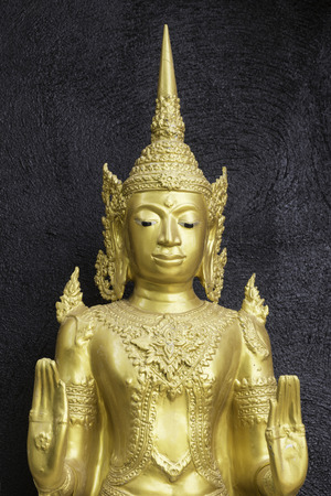 Yellow statue wearing a Thai crown on a backdrop of black painted plaster at a temple in Thailand.の写真素材