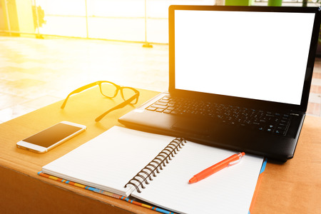 Blank screen of laptop with pen on notebook and cell phone on a wooden table in sunny room, soft...