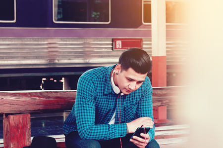 Handsome young asian man looking smart phone and play music with waitting train at station.の写真素材