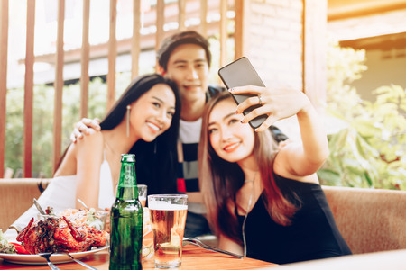Group of asian friends sitting at bar restaurant and taking selfie with mobile smartphone.の写真素材