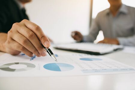 Close up hand of a businessman holding a pen pointing to graph of financial statements or company profits and analyzing on the desk at the office room.の写真素材
