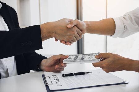 Two corporate businessmen shaking hands while one man places money on document in office room with...