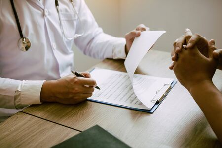 Doctor hand holding pen writing patient history list on note pad and talking to the patient about...