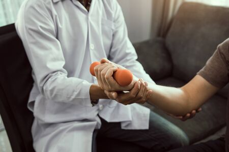 Asian young male physiotherapist helping patient with lifting dumbbells exercises in office.