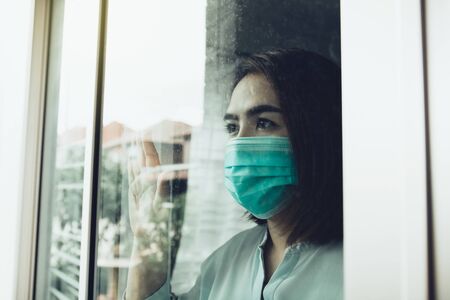 Asian woman looking through the window and wearing medical mask protection from the illness with...