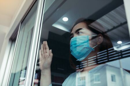 Asian woman looking through the window and wearing medical mask protection from the illness with...