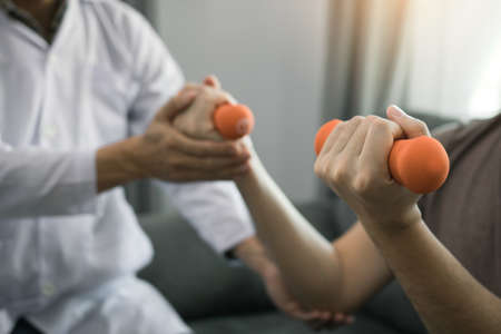 Asian physiotherapist helping a patient lifting dumbbells work through his recovery with weights in clinic room.の写真素材