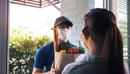 Delivery of asian man wearing a protective mask during an epidemic virus while he was delivering a bag of vegetable food to a female customer at the door.の写真素材