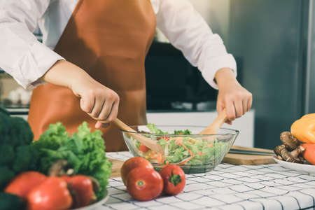 Asian woman is mixing the ingredients in a salad bowl at the kitchen cooking table.の写真素材