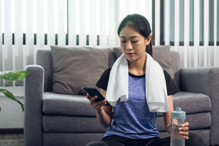 Asian woman playing smartphone at home during break from yoga exercise and drinking fresh water.