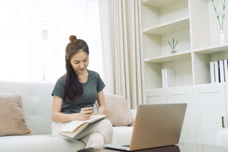 Asian woman sits on the sofa while taking notes from her computer screen while working or studying...