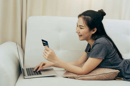 Asian woman holding a credit card for online shopping while sitting on the couch while on vacation at home.の写真素材