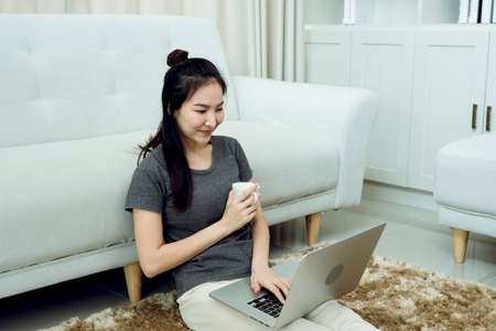 Asian woman is sitting in the living room holding a coffee cup and a laptop computer on her lap at...