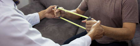 Asian male physical therapist descent working and helping to protect the hands of patients with patient doing stretching exercise with a flexible exercise band in clinic room.の写真素材