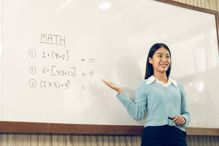 Asian female teacher is teaching students at the classroom while pointing at numbers on the white board.の写真素材