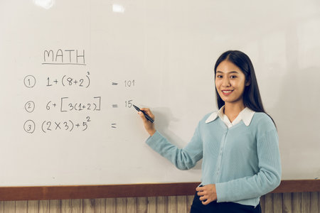 Asian female teacher is teaching students at the classroom while pointing at numbers on the white board.の写真素材