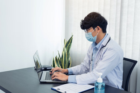 Doctor was sitting in the office working on a laptop computer while wearing a mask.の写真素材