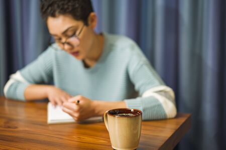 A woman wearing glasses sits at a table drinking coffee and writing in a journalの写真素材