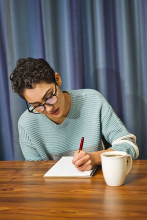 A woman wearing glasses sits at a table drinking coffee and writing in a journalの写真素材