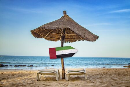 United Arab Emirates flag on wooden arrow sign. There are two sun loungers and a sun umbrella on the beach. It is a tropical paradise with a clear sea. It's summer vacation time.の写真素材