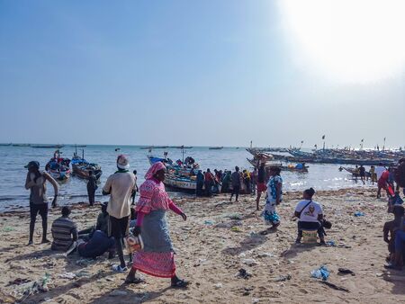 MBour, Senegal- April 25 2019: Unidentified Senegalese men and women at the fish market in the port city near Dakar. There are stalls selling and fishing boats everywhere. It is sunny day.のeditorial素材