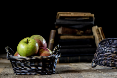 Tasty apples in basket on kitchen table. Old books lie next to apples in a garbage. Autumn season. Wooden table, black backgroundの写真素材