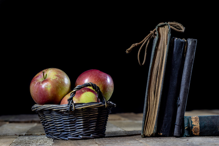 Tasty apples in basket on kitchen table. Old books lie next to apples in a garbage. Autumn season. Wooden table, black backgroundの写真素材