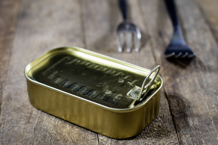 A tin can of fish on a wooden kitchen table. Next to the can fork. Preserved fish in a tin. Old...