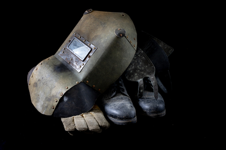 An old mask a protective suit for a welder. Occupational health and safety clothing for production workers on a workshop table. Black background.の写真素材