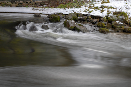 A small river flowing between trees with a small water degree. The riverbed leading through a deciduous forest. Season winter.の写真素材