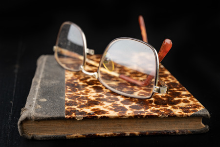 Old book and glasses on a dark wooden table. Forgotten publishing in binding from several decades ago. Black background.の写真素材