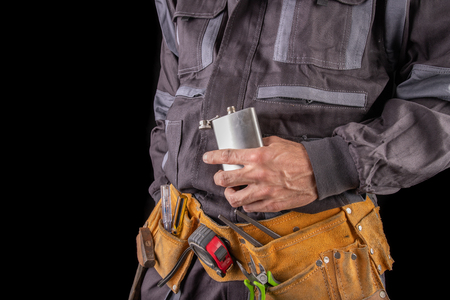 A worker in protective clothing and a flask with vodka. Drunk production worker in the workplace. Dark background.の写真素材