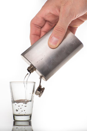 Pouring high-percentage alcohol into a glass shot glass. Metal flask with alcohol and a glass shot glass. Light background.の写真素材