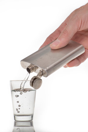 Pouring high-percentage alcohol into a glass shot glass. Metal flask with alcohol and a glass shot glass. Light background.の写真素材
