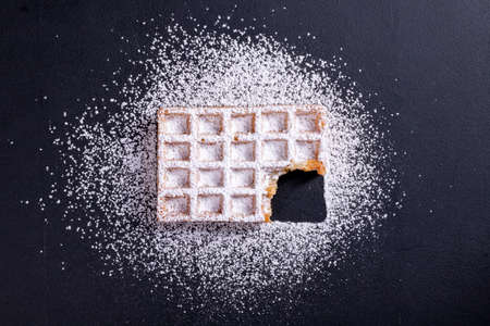 Sweet waffles sprinkled with powdered sugar prepared for serving. A tasty dessert eaten with fruit and preserves. Dark background.の写真素材