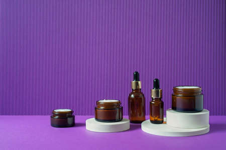 Amber glass bottles and jars with trendy geometric shapes on a violet background. White pedestals . Skincare products , natural cosmetic. Beauty concept for face and body careの写真素材