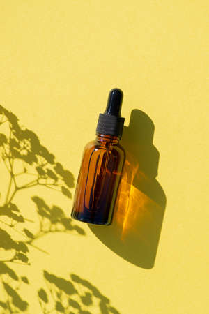 Amber glass dropper bottle with black lid. Top view on yellow background with plant shadow. Skincare...