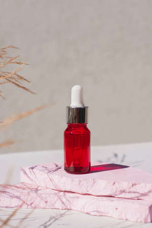 Red glass dropper bottle with silver lid on a podium with dry plants on light background. Skincare products , natural cosmetic. Beauty concept for face and body careの写真素材