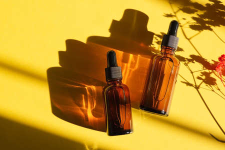 Amber glass dropper bottles with black lid. Top view on yellow background with plant shadow....