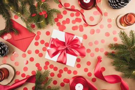 White gift box with red ribbon with red confetti on a kraft background. Packaging and preparation of gifts for the celebration. Christmas New Year backdrop with tree branch decor and lightsの写真素材