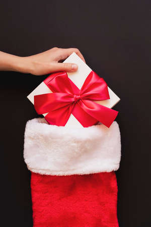 Womans hand puts a white gift with a red bow in red Santas socks. Packaging and preparation of gifts...