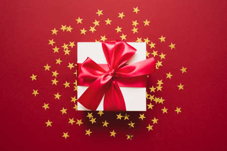 White gift box with ribbon on a red with golden confetti in the form of stars. Valentines, Christmas...