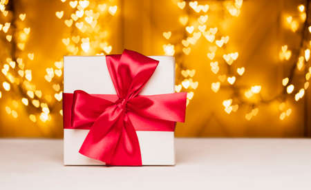 White gift box with ribbon on a red against the background of bokeh lights in the form of hearts. Valentines, Christmas or New Year day backdrop.の写真素材